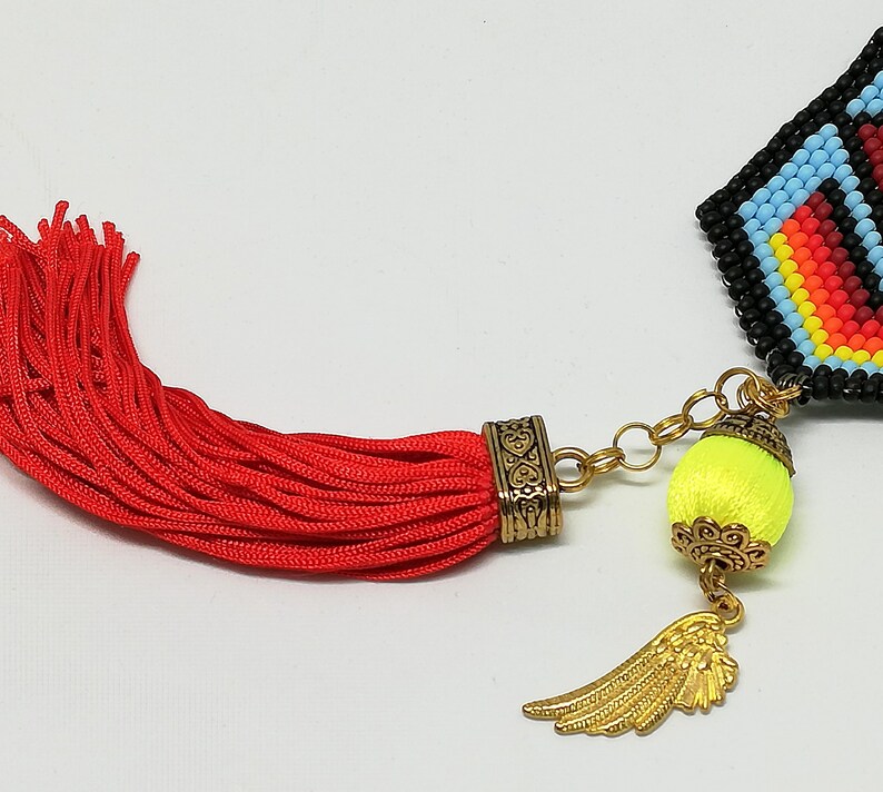 May include: Close-up of a beaded pendant with a red tassel. The pendant features a geometric design in black, blue, red, and yellow. A gold-colored wing charm and a neon yellow ball are attached.