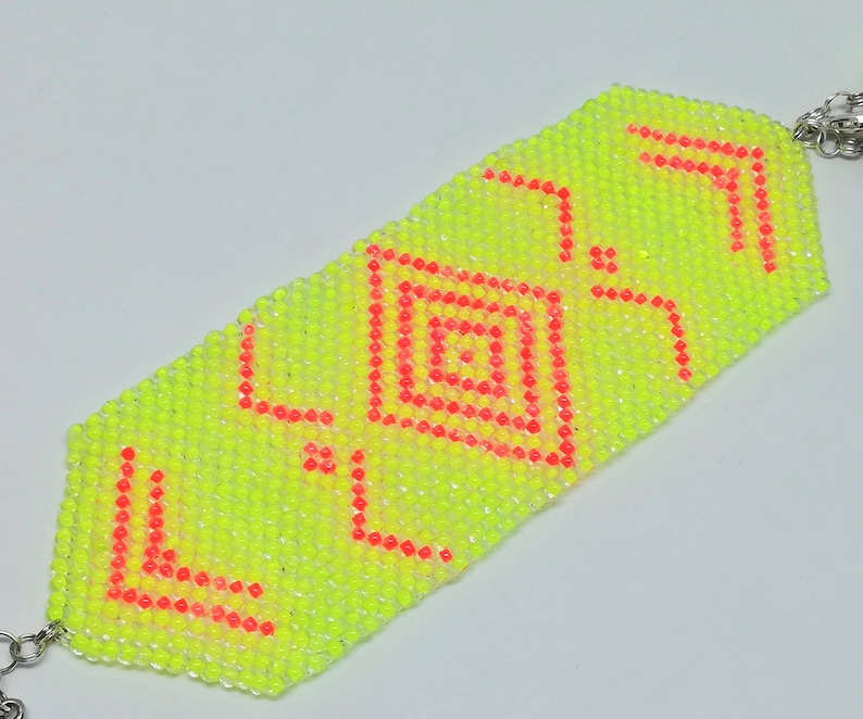 May include: A neon yellow beaded bracelet with a geometric design in red. The bracelet features a central square pattern and angular accents, connected by silver-coloured chain links.