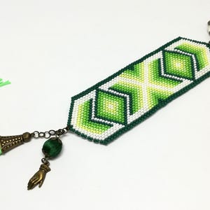 May include: A beaded bracelet with a geometric design in shades of green and white. The bracelet features a green tassel and decorative charms, including a hand and a green bead. The bracelet is approximately 7 inches long.