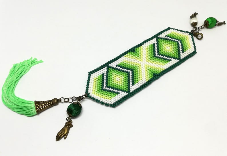 May include: A beaded bracelet with a geometric design in shades of green and white. The bracelet features a green tassel and decorative charms, including a hand and a green bead. The bracelet is approximately 7 inches long.