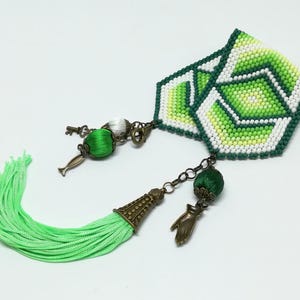 May include: A handmade bookmark with a geometric design in shades of green, white, and yellow. It features a green tassel and decorative charms, including a key and a hand. The bookmark is designed for book lovers.