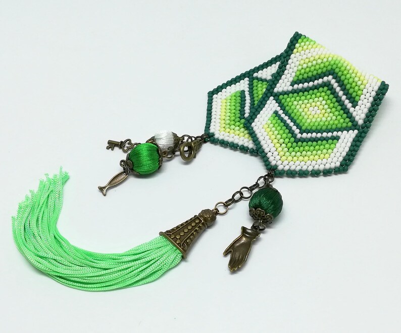 May include: A handmade bookmark with a geometric design in shades of green, white, and yellow. It features a green tassel and decorative charms, including a key and a hand. The bookmark is designed for book lovers.