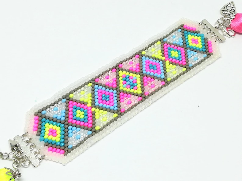 May include: A colorful beaded bracelet featuring a geometric pattern of pink, yellow, blue, and gray squares. The bracelet has a white base and silver-tone clasps. A pink tassel and charm dangle from the end.