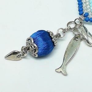 May include: Close-up of a blue beaded charm with a silver-toned fish-shaped charm and a decorative blue silk-wrapped bead. The charm is attached to a silver chain and clasp, with a white background.