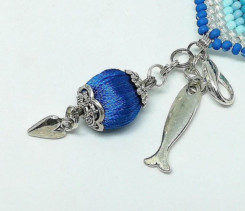 May include: Close-up of a blue beaded charm with a silver-toned fish-shaped charm and a decorative blue silk-wrapped bead. The charm is attached to a silver chain and clasp, with a white background.
