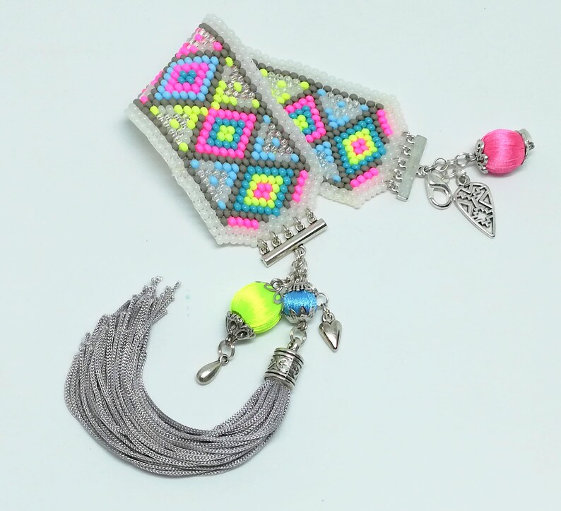 May include: A colorful beaded bracelet with a geometric pattern in neon pink, yellow, blue, and gray. The bracelet has silver-tone clasps and charms, including a tassel and a pink silk-wrapped bead. The bracelet is a unique fashion accessory.