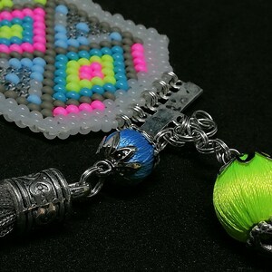 May include: Close-up of a beaded accessory with a geometric pattern in pink, yellow, blue, and gray. The accessory features a silver tassel and a bright green orb. The design includes silver chain links and a blue bead.