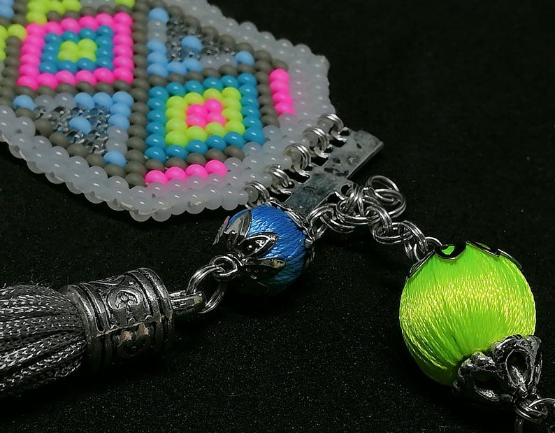 May include: Close-up of a beaded accessory with a geometric pattern in pink, yellow, blue, and gray. The accessory features a silver tassel and a bright green orb. The design includes silver chain links and a blue bead.