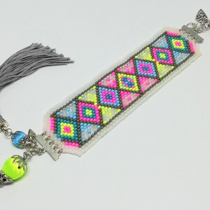May include: A handmade bookmark with a colorful geometric pattern. The design features pink, blue, and yellow squares and triangles on a white background. It has a silver tassel and decorative beads. The bookmark is a unique accessory for book lovers.
