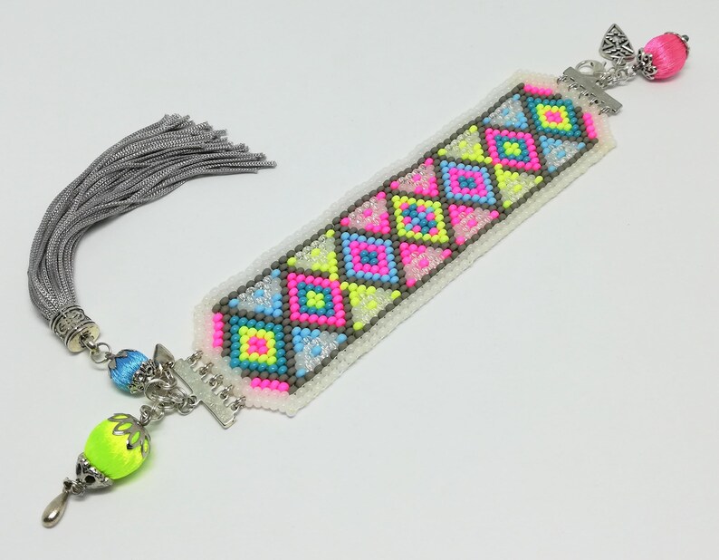 May include: A handmade bookmark with a colorful geometric pattern. The design features pink, blue, and yellow squares and triangles on a white background. It has a silver tassel and decorative beads. The bookmark is a unique accessory for book lovers.