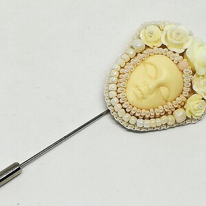 May include: A white and cream beaded brooch with a sculpted face and white flower accents. The brooch is attached to a silver pin.