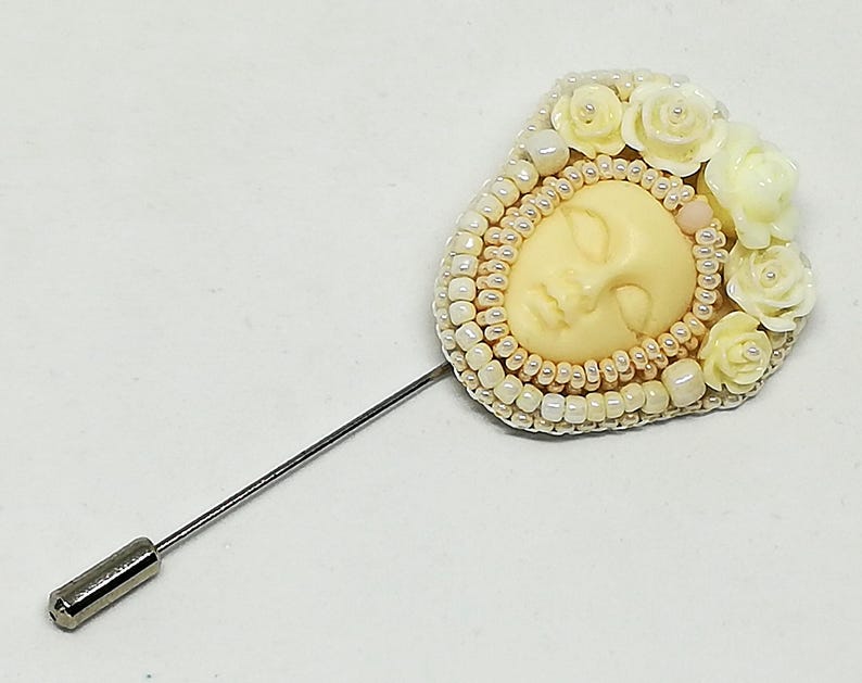 May include: A white and cream beaded brooch with a sculpted face and white flower accents. The brooch is attached to a silver pin.