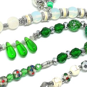 May include: Close-up of beaded jewelry strands. The strands feature a variety of beads in white, green, and silver. Some beads are faceted, others are round, and some are teardrop-shaped. The jewelry has a handmade appearance.