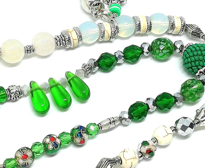 May include: Close-up of beaded jewelry strands. The strands feature a variety of beads in white, green, and silver. Some beads are faceted, others are round, and some are teardrop-shaped. The jewelry has a handmade appearance.
