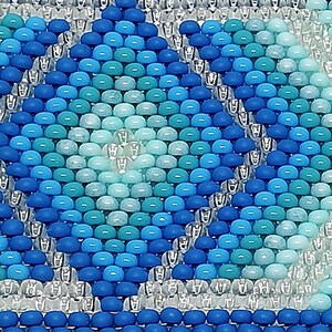 May include: Close-up of a beaded pattern in shades of blue and turquoise. The design features diamond shapes and a border of clear beads. The beads are small and evenly spaced, creating a textured surface. The overall effect is a mosaic-like appearance.
