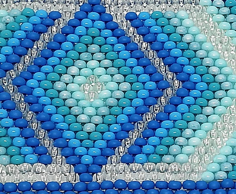 May include: Close-up of a beaded pattern in shades of blue and turquoise. The design features diamond shapes and a border of clear beads. The beads are small and evenly spaced, creating a textured surface. The overall effect is a mosaic-like appearance.