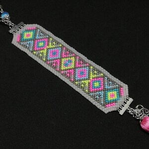 May include: Handmade beaded bracelet with a geometric pattern in pink, yellow, blue, and gray. The bracelet has silver-tone metal accents and a pink tassel. A unique, colorful accessory.