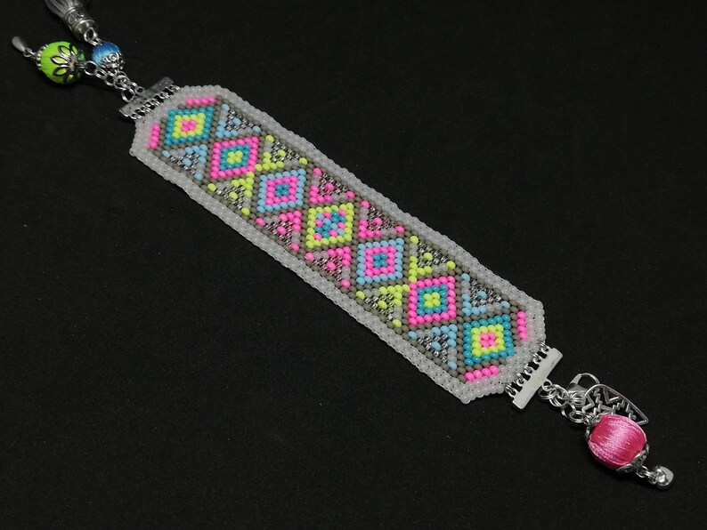 May include: Handmade beaded bracelet with a geometric pattern in pink, yellow, blue, and gray. The bracelet has silver-tone metal accents and a pink tassel. A unique, colorful accessory.