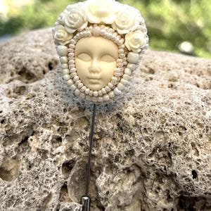 May include: A white porcelain face with closed eyes, surrounded by white pearl beads and white roses, is attached to a silver pin.