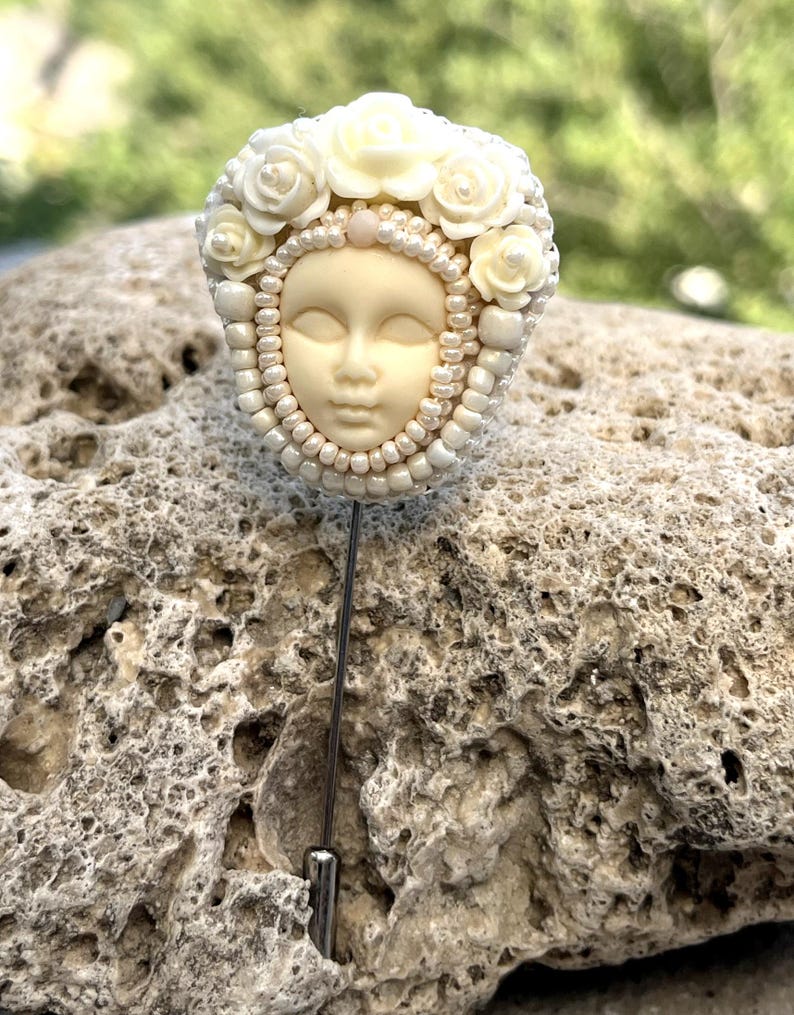 May include: A white porcelain face with closed eyes, surrounded by white pearl beads and white roses, is attached to a silver pin.