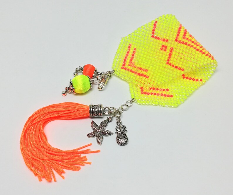 May include: A neon yellow beaded accessory with orange detailing. Features an orange tassel, silver charms of a starfish and pineapple, and a beaded geometric design. Includes a chain and two small beaded spheres.