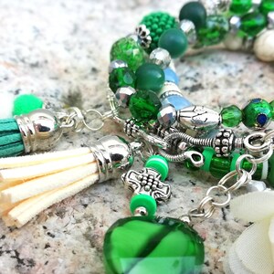 May include: Close-up of a handmade bracelet with green beads, silver accents, and a green tassel. The bracelet features various shades of green beads, including round, faceted, and textured beads. The tassel is made of cream-colored fringe and green suede.