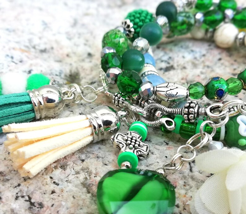 May include: Close-up of a handmade bracelet with green beads, silver accents, and a green tassel. The bracelet features various shades of green beads, including round, faceted, and textured beads. The tassel is made of cream-colored fringe and green suede.
