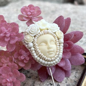 May include: A white porcelain face with closed eyes surrounded by white flowers and pearls, attached to a pin.