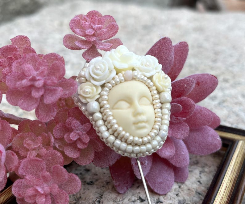 May include: A white porcelain face with closed eyes surrounded by white flowers and pearls, attached to a pin.