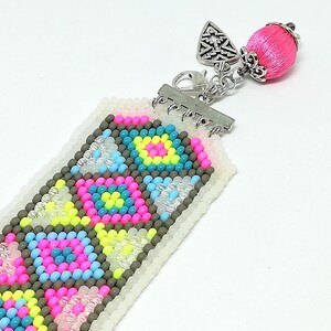May include: A beaded bookmark with a geometric pattern in pink, blue, yellow, and gray. The bookmark has a silver clasp and a pink tassel with a silver cap. The beads are small and tightly woven together, creating a detailed design.