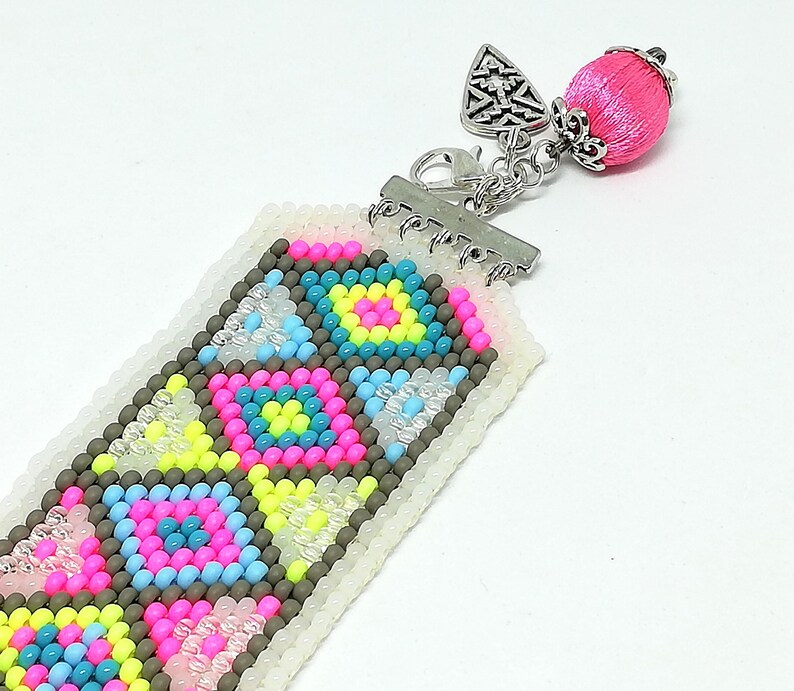 May include: A beaded bookmark with a geometric pattern in pink, blue, yellow, and gray. The bookmark has a silver clasp and a pink tassel with a silver cap. The beads are small and tightly woven together, creating a detailed design.
