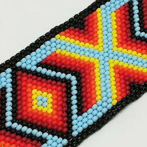 May include: Close-up of a beaded bracelet with a geometric pattern. The design features a central cross-like shape in red, orange, and yellow, bordered by light blue and black beads. The bracelet is made of small, round beads.