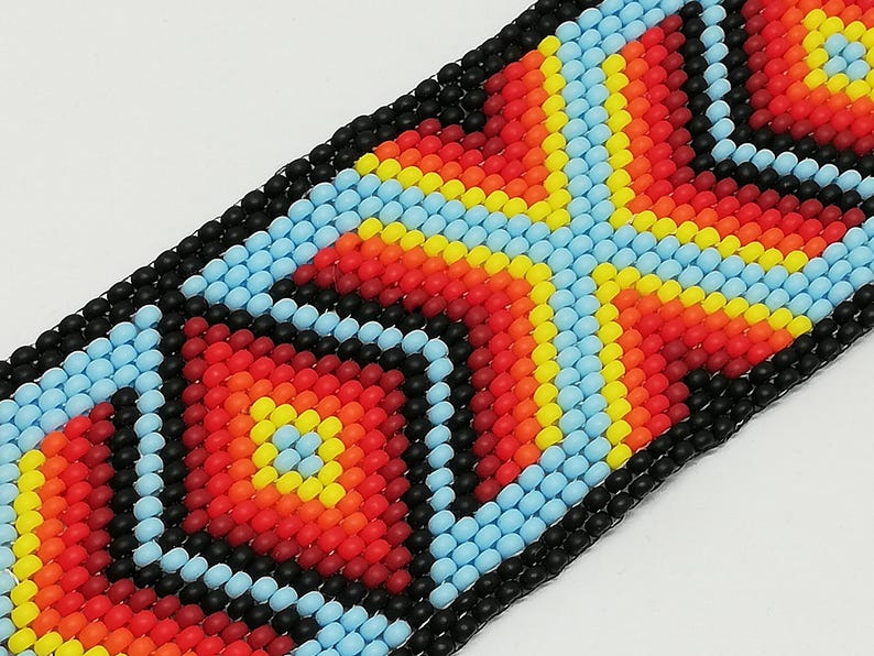 May include: Close-up of a beaded bracelet with a geometric pattern. The design features a central cross-like shape in red, orange, and yellow, bordered by light blue and black beads. The bracelet is made of small, round beads.
