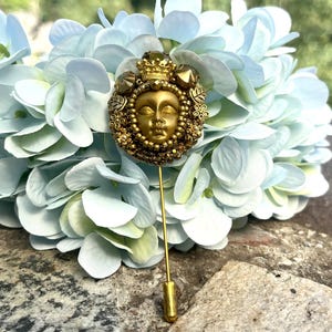 May include: A gold-toned stick pin featuring a detailed face with a crown and ornate embellishments. The pin is set against a backdrop of light blue and white flowers, creating a striking contrast. The pin's design suggests a vintage or antique style.