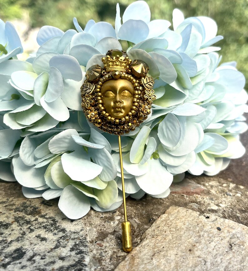 May include: A gold-toned stick pin featuring a detailed face with a crown and ornate embellishments. The pin is set against a backdrop of light blue and white flowers, creating a striking contrast. The pin's design suggests a vintage or antique style.