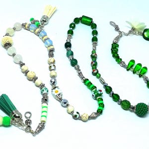 May include: Three beaded necklaces with various designs. One features white, cream, and green beads with a green tassel. Another is primarily green with silver accents. The third has green beads and a flower charm.