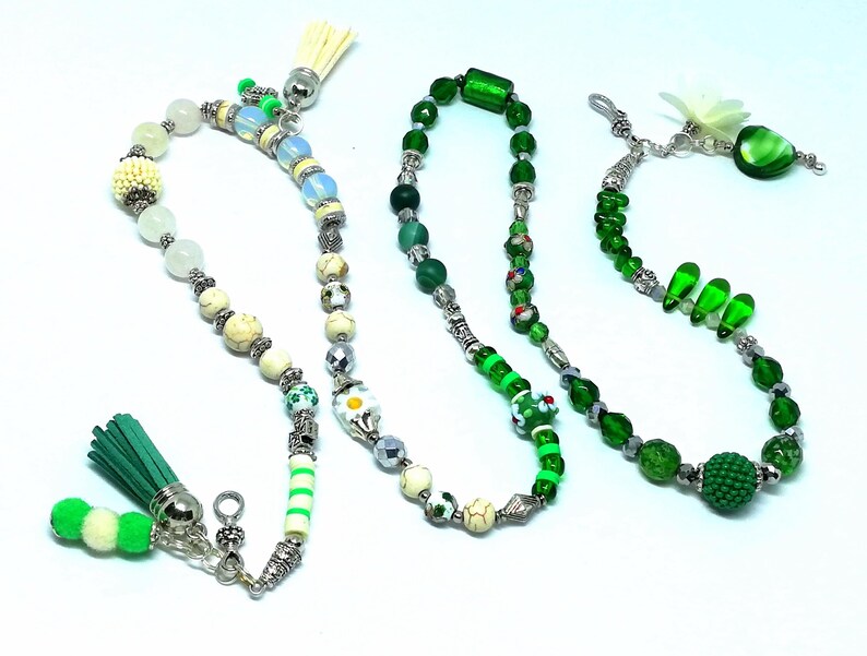 May include: Three beaded necklaces with various designs. One features white, cream, and green beads with a green tassel. Another is primarily green with silver accents. The third has green beads and a flower charm.