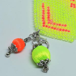 May include: A close-up of a keychain featuring two decorative silk thread balls in neon orange and green, each with a silver filigree cap and a small silver bead. The keychain is attached to a beaded neon yellow and red square.