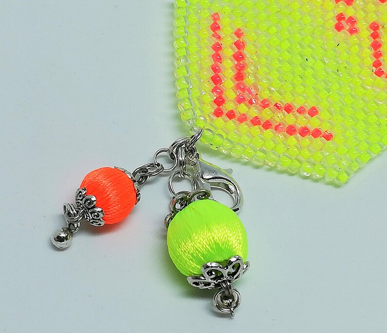 May include: A close-up of a keychain featuring two decorative silk thread balls in neon orange and green, each with a silver filigree cap and a small silver bead. The keychain is attached to a beaded neon yellow and red square.