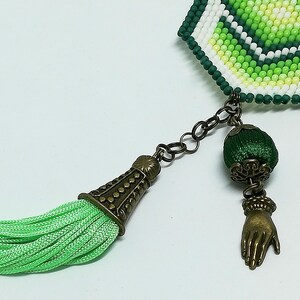 May include: A beaded bookmark with green, white, and yellow beads. It features a green tassel, a dark metal chain, and a charm of a hand. The bookmark has a decorative metal end cap.