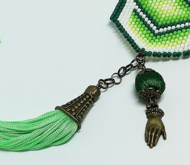 May include: A beaded bookmark with green, white, and yellow beads. It features a green tassel, a dark metal chain, and a charm of a hand. The bookmark has a decorative metal end cap.