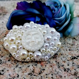 May include: A white pearl brooch with a white face in the center. The face has closed eyes and a small mouth.