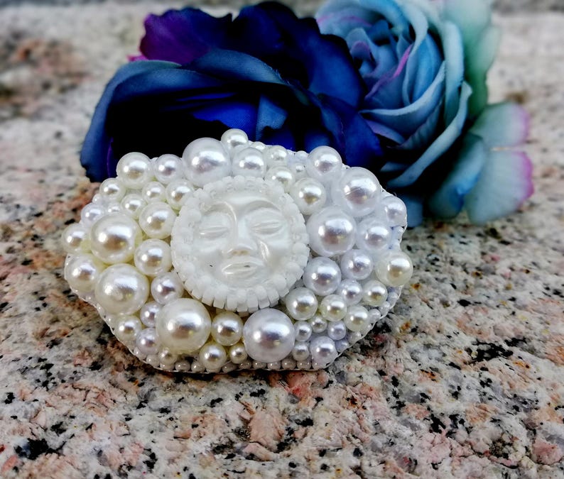 May include: A white pearl brooch with a white face in the center. The face has closed eyes and a small mouth.