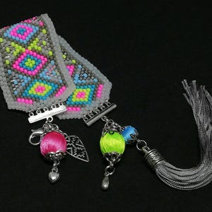 May include: A beaded bracelet with a geometric pattern in pink, blue, yellow, and gray. The bracelet has silver clasps and is accompanied by a pink and green beaded charm, a silver charm, and a gray tassel.