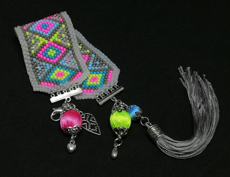 May include: A beaded bracelet with a geometric pattern in pink, blue, yellow, and gray. The bracelet has silver clasps and is accompanied by a pink and green beaded charm, a silver charm, and a gray tassel.