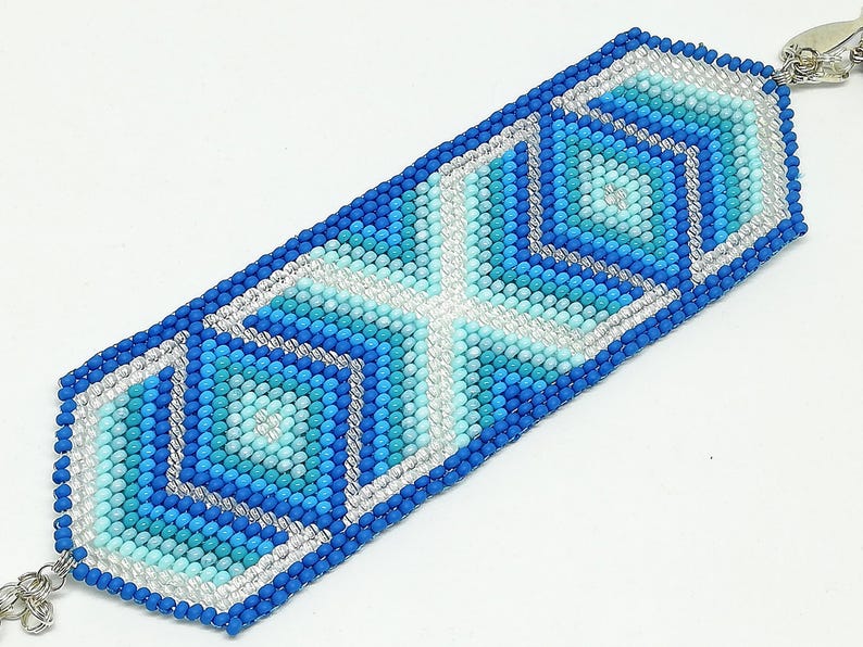 May include: A beaded bracelet featuring a geometric design in shades of blue, turquoise, and white. The bracelet is rectangular with a silver clasp and a detailed pattern of squares and diamonds.