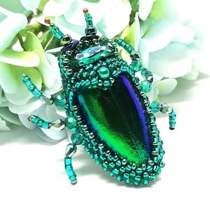 Op de afbeelding: Een broche in de vorm van een groene en blauwe iriserende kever gemaakt van kralen. De kever is versierd met iriserende groene en blauwe kralen en heeft een glanzende afwerking.