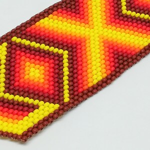 Op de afbeelding: Close-up van een kralenarmband met een geometrisch ontwerp. De armband heeft een patroon van gele, oranje en rode kralen, met een bruine rand. De kralen zijn klein en rond en cre&euml;ren een textuur. Het ontwerp is symmetrisch en opvallend.