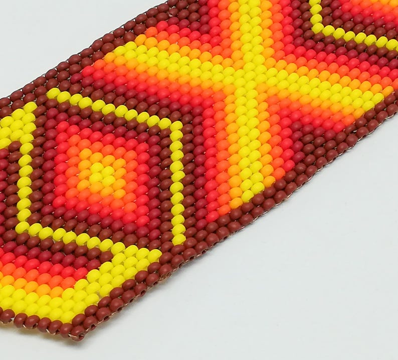Op de afbeelding: Close-up van een kralenarmband met een geometrisch ontwerp. De armband heeft een patroon van gele, oranje en rode kralen, met een bruine rand. De kralen zijn klein en rond en cre&euml;ren een textuur. Het ontwerp is symmetrisch en opvallend.