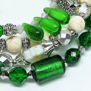May include: Close-up of beaded jewelry featuring a mix of green, silver, and cream-colored beads. The beads include faceted green crystals, cylindrical green glass beads, and round cream-colored beads with a marbled effect. The jewelry is strung on a clear cord.